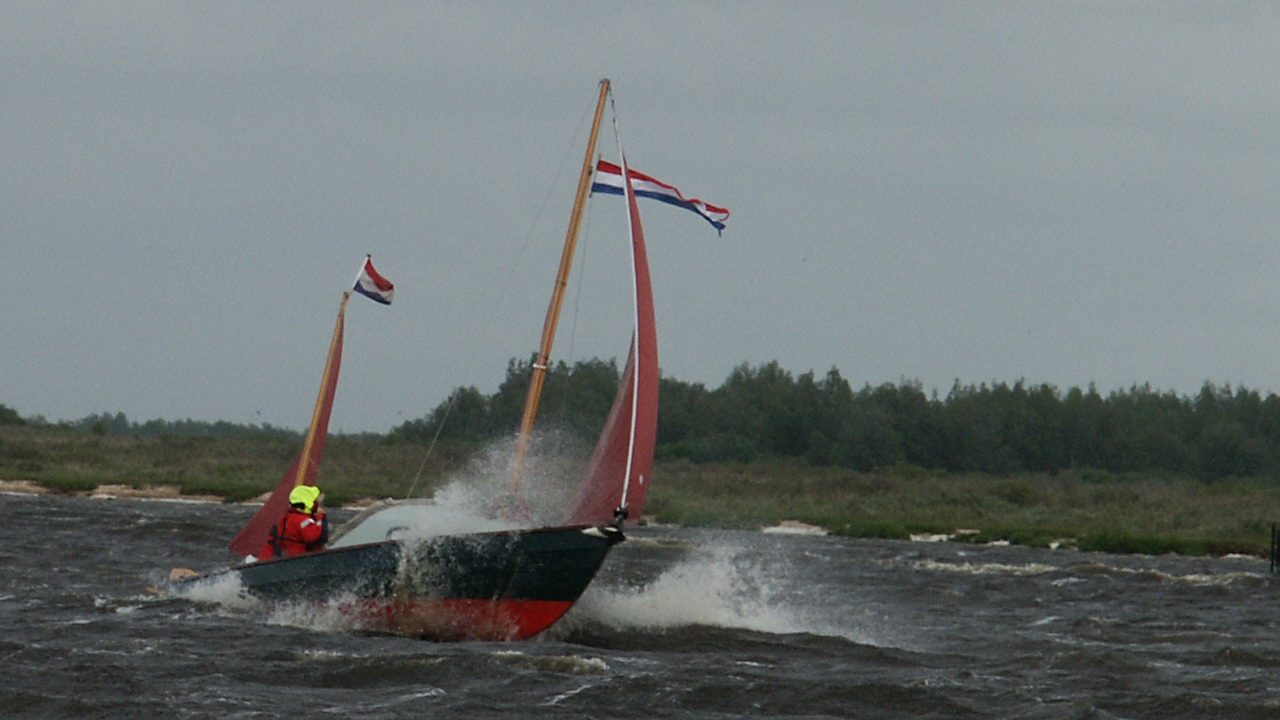 Een Coaster met roodbruine zeilen en Nederlandse vlaggen zeilt hard door de golven, met opspattend water langs de boeg.